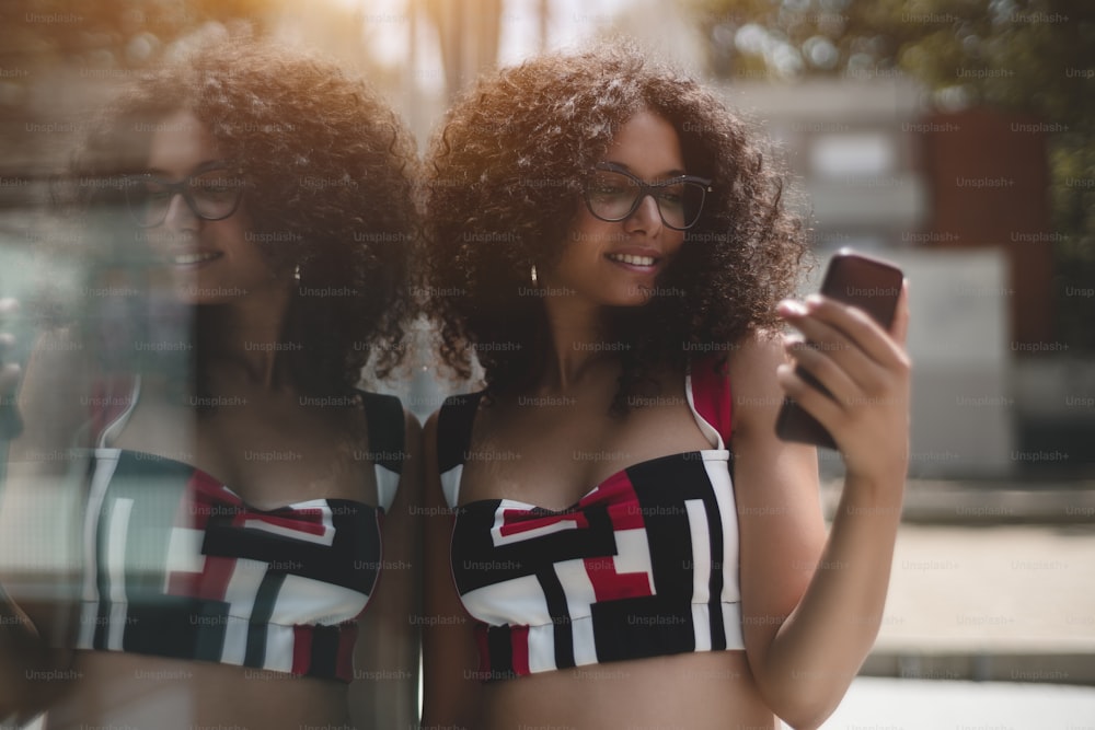 Portrait of charming caucasian girl in eyeglasses and with bulky curly hair, she is leaning against the glass wall outdoors which fully reflects her and looking at the screen of her smartphone