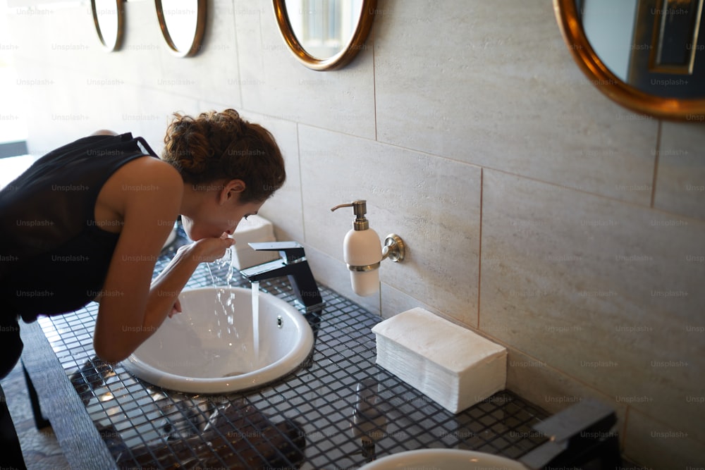 Junge Frau wäscht sich den Mund mit Wasser aus dem Wasserhahn, während sie sich über das Waschbecken in der Toilette beugt
