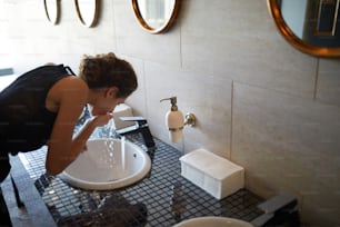 Young woman washing her mouth with water from tap while leaning over sink in lavatory