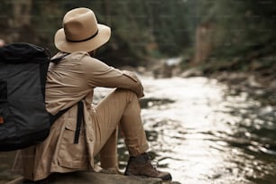 깊은 생각. 배낭을 메고 모자를 쓰고 여행하는 동안 강 근처에 앉아 있는 청년