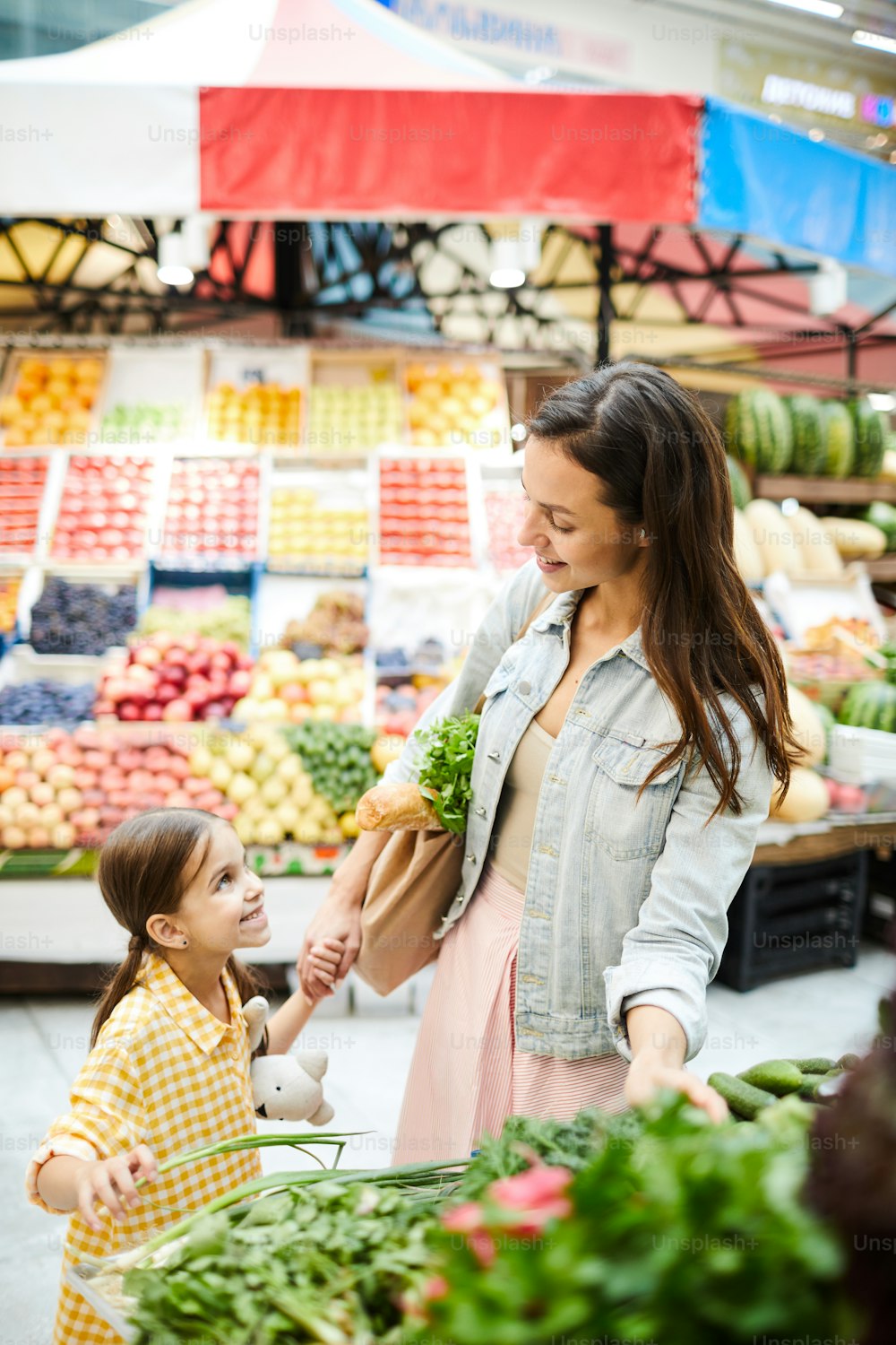 캐주얼 재킷을 입은 매력적인 젊은 어머니가 신선한 음식 가판대에 서서 식품점에서 딸과 이야기하면서 야채를 고르고 있다