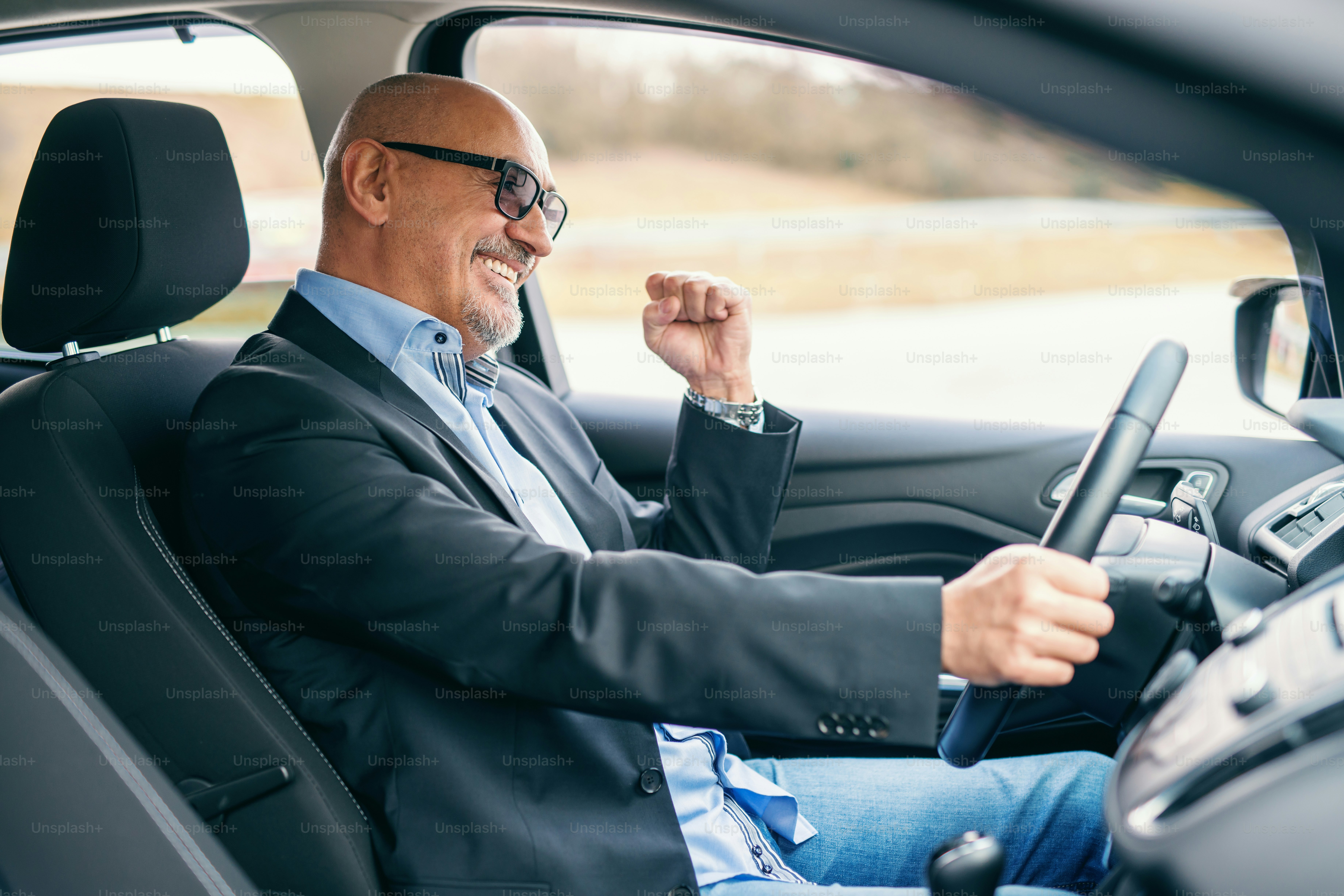 Bearded smiling senior adult businessman driving car during the day. Hand on the wheel.