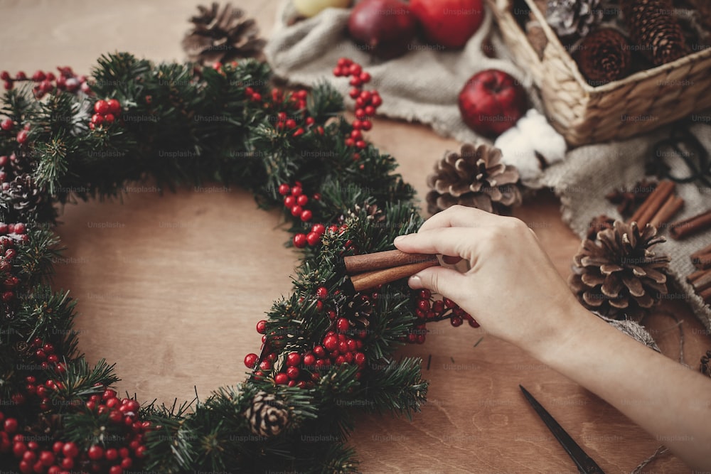 素朴な木製の背景にモミの枝、赤い果実、松ぼっくり、ロープ、はさみ、綿でシナモンを保持し、素朴なクリスマスリースを作る手。休日のワークショップでの雰囲気のある不機嫌なイメージ