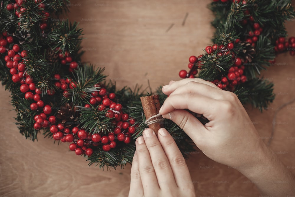 Hands making rustic christmas wreath, holding cinnamon at fir branches, red berries , pine cones, cotton on rustic wooden background. Atmospheric moody image at winter holiday workshop