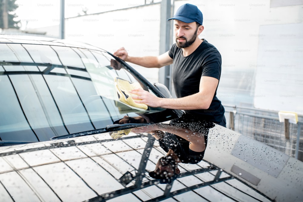 Guapo lavaparabrisas en camiseta y gorra limpiando el parabrisas con microfibra amarilla en el lavado de autos al aire libre