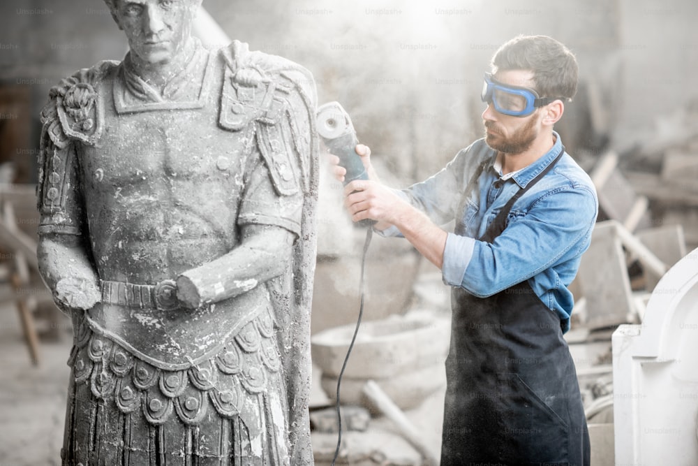 埃のある古いスタジオで電気グラインダーで石の彫刻を砥石する保護作業器具の彫刻家