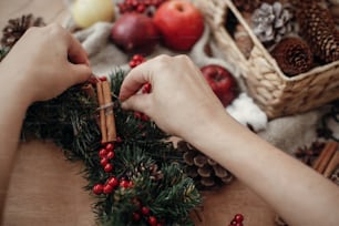 Hands making rustic christmas wreath, holding cinnamon at fir branches, red berries,pine cones,rope, scissors, cotton on rustic wooden background. Atmospheric moody image at holiday workshop