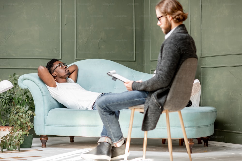 Young afro ethnicity man having a serious conversation with psychologist lying on the comfortable couch during psychological session in the office