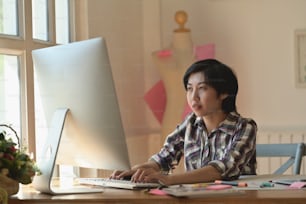 Stylish Asian fashion designer working with modern desktop computer in atelier.