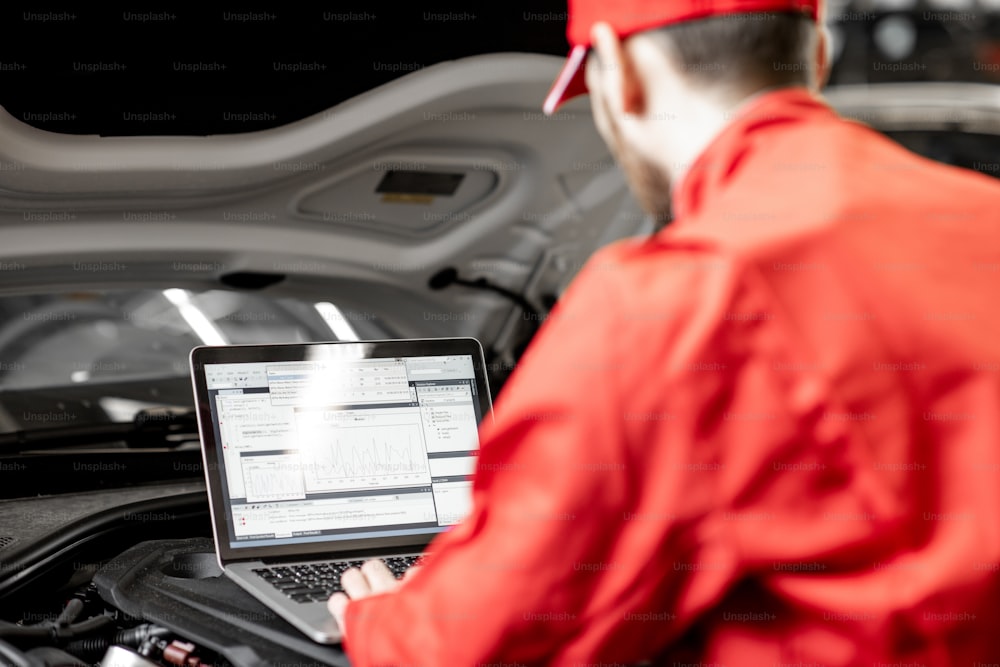 赤い制服を着たハンサムな自動車整備士が、車のサービスでコンピューターを使用してエンジン診断を行っています