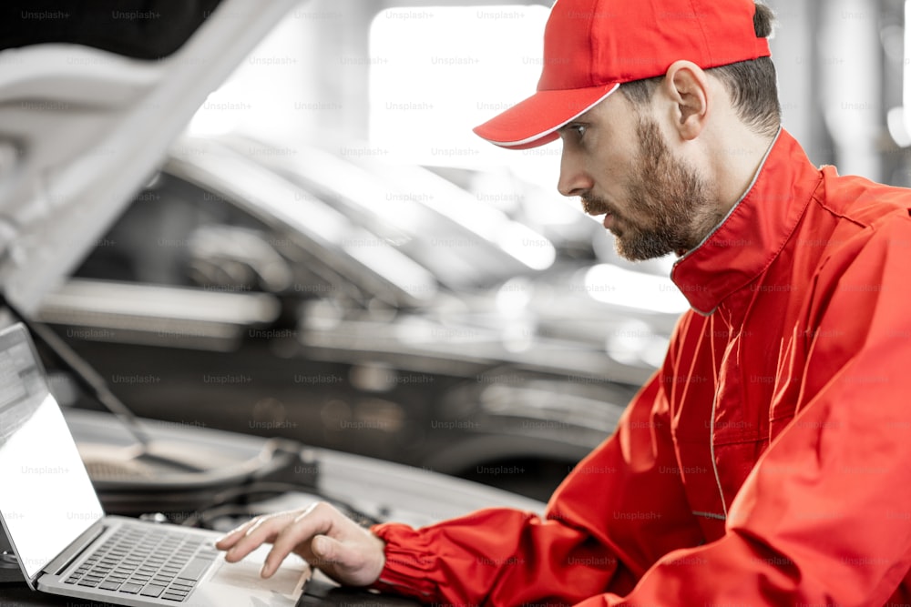Meccanico d'auto bello in uniforme rossa che fa la diagnostica del motore con il computer nel servizio dell'automobile