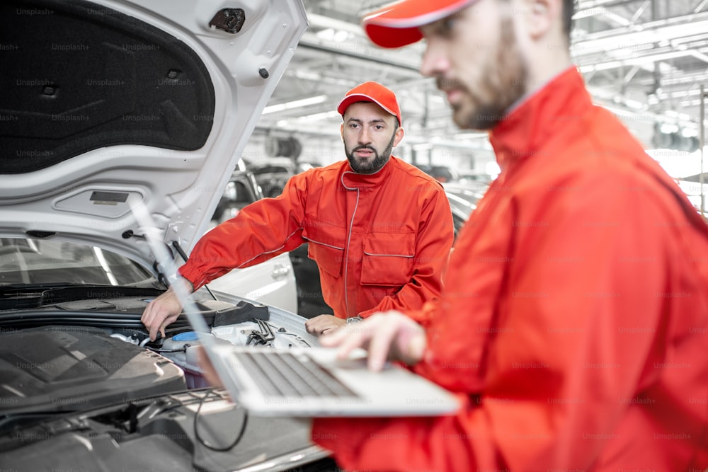 Two auto mechanics in red uniform doing engine diagnostics with computer in the car service