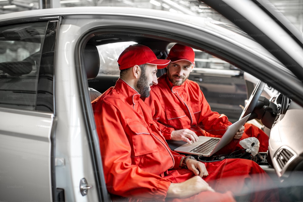 Two male auto mechanics in red uniform diagnosing car with computer sitting on the seats indoors at the car service