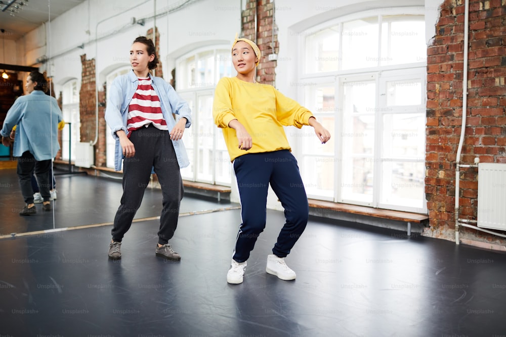 Two young active women doing hip hop exercises on the floor with large mirror on background