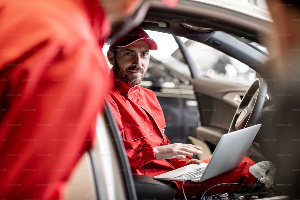 Auto mechanic in red uniform diagnosing car with computer sitting on the driver seat at the car service