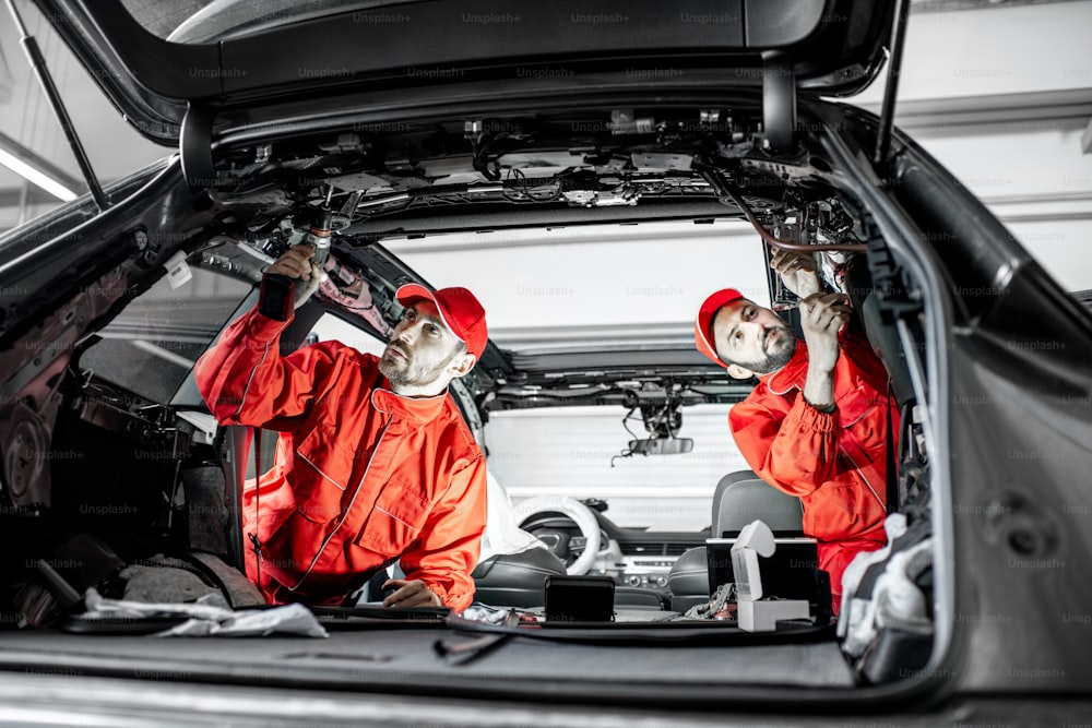 Two auto service workers in red uniform disassembling new car interior making some improvements indoors