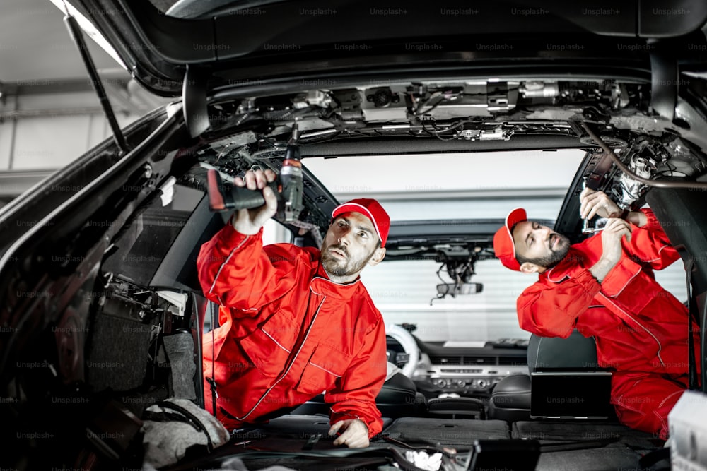 Two auto service workers in red uniform disassembling new car interior making some improvements indoors
