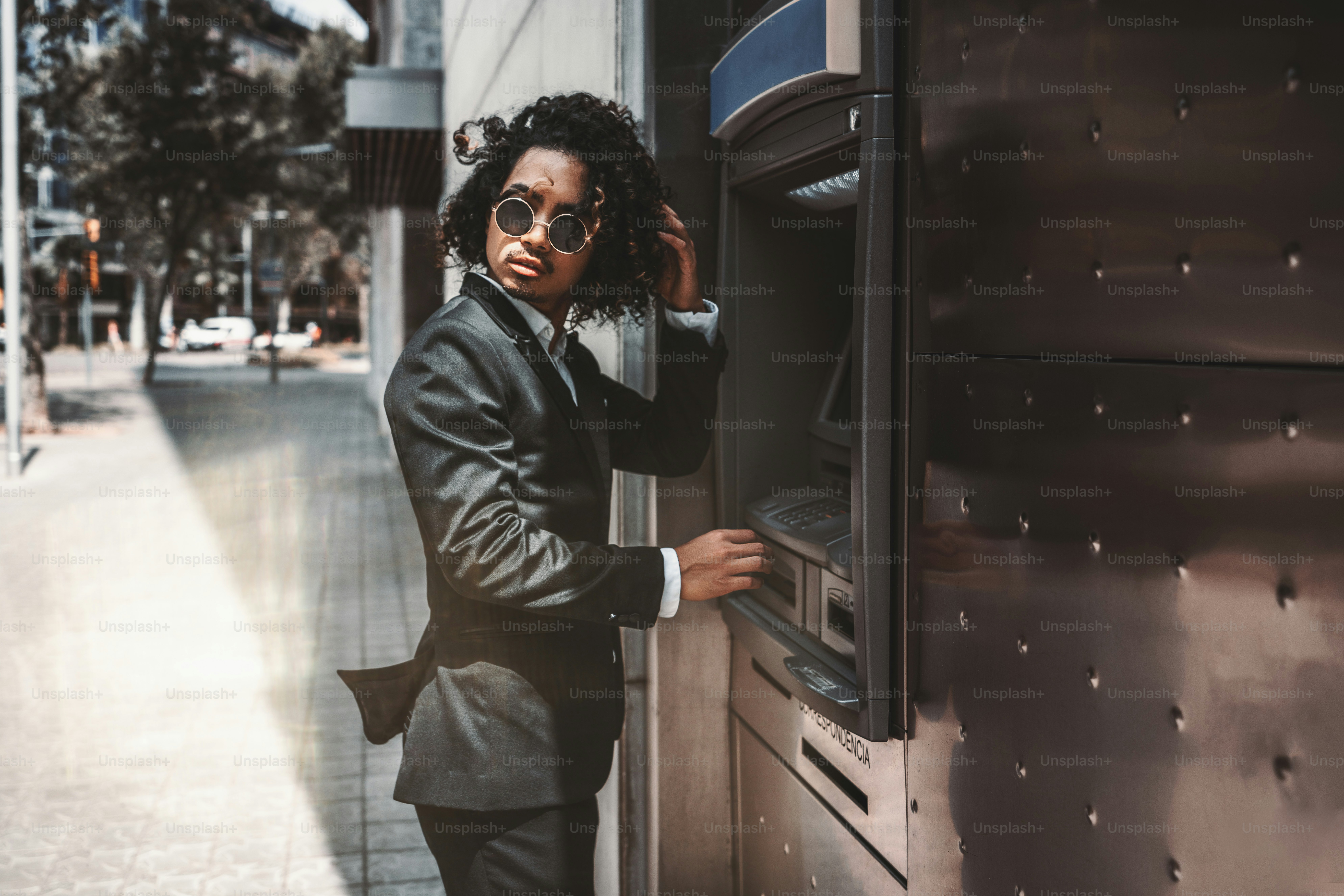 An Asian curly man employer in sunglasses and business suit is looking aside and fixing his hair while standing near an outdoor ATM; businessman on the street withdrawing money via the cash dispenser