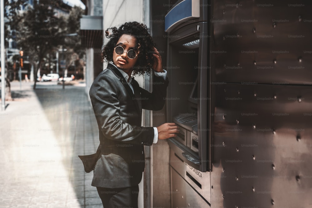 An Asian curly man employer in sunglasses and business suit is looking aside and fixing his hair while standing near an outdoor ATM; businessman on the street withdrawing money via the cash dispenser