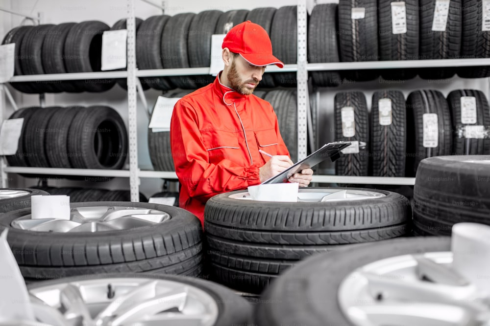Worker or salesman in red uniform filling some documents checking goods in the warehouse with car tires