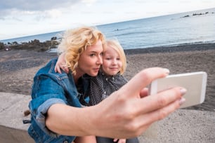 ブロンドの女性と小さなブロンドの息子が海と地平線を背景に自撮り写真を撮ります。新しいテクノロジーで休暇のライフスタイルの思い出。屋外の若い家族の幸せ