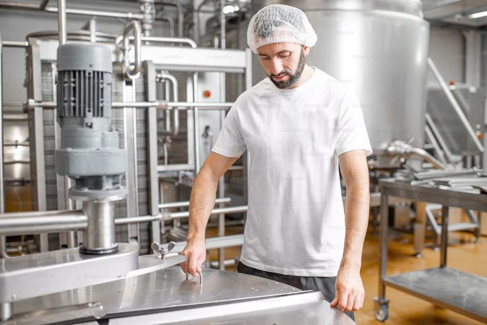 Retrato de un apuesto trabajador en uniforme cerca del tanque de acero inoxidable lleno de leche fermentada en la fabricación de queso