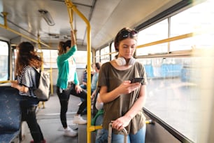Beautiful Caucasian girl with brown hair and headphones around neck using smart phone for reading or sending message while standing in public transportation.
