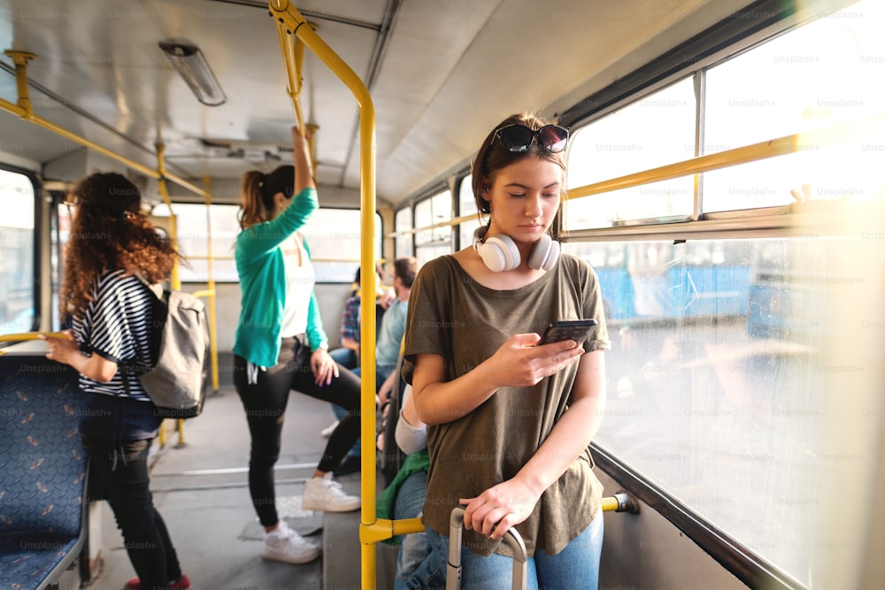갈색 머리와 헤드폰을 목에 두른 아름다운 백인 소녀가 대중 교통에 서 있는 동안 스마트폰을 사용하여 메시지를 읽거나 보냅니다.