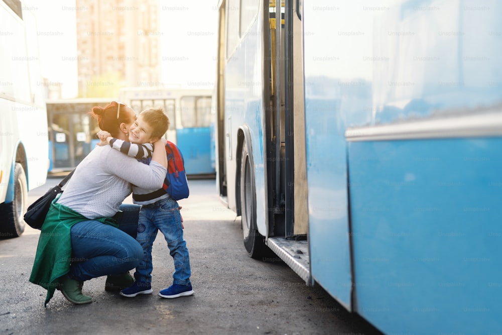 Mother hugging her son while crouching next to bus. Boy going to school.