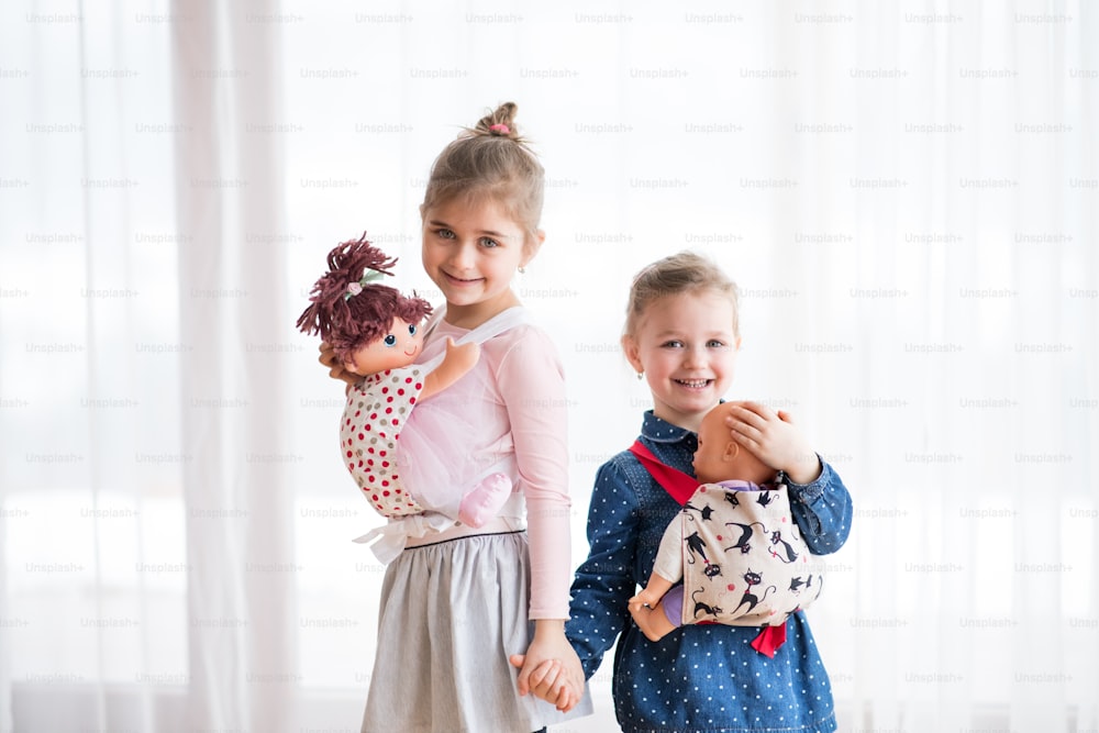 A portrait of two small girls standing and carrying dolls in baby carriers indoors, holding hands.