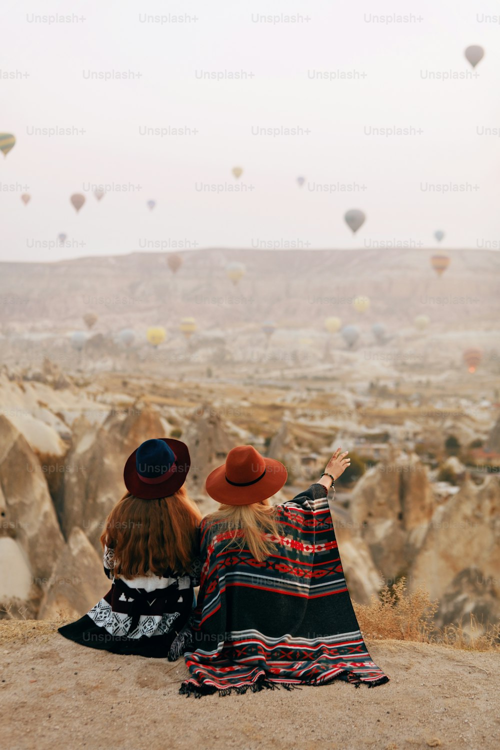 人は旅をします。帽子をかぶった女性が丘の上に座り、トルコのカッパドキアで熱気球を飛ばす様子を楽しんでいます。高解像度