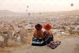La gente viaja. Mujeres En Sombreros Sentadas En La Colina Disfrutando De Volar Globos Aerostáticos Vista En Capadocia, Turquía. Alta resolución