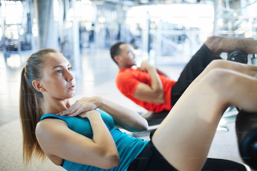 Young sportswoman and her mate on background doing sit-ups on the floor during workout in gym