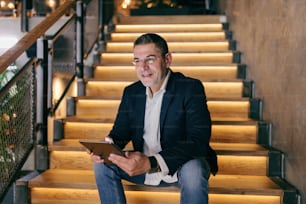 Homme d’affaires caucasien souriant d’âge moyen, habillé de manière élégante et décontractée, assis dans les escaliers d’un café tout en utilisant une tablette.