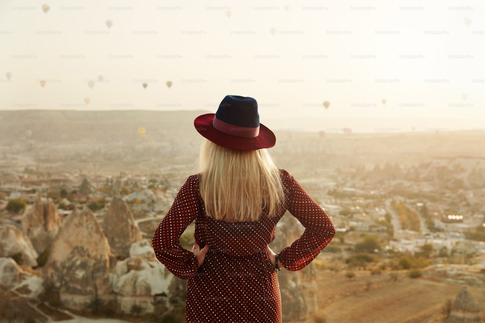 旅行。空に飛ぶ熱気球と丘の上の帽子をかぶった美しい女性。カッパドキアに旅行する女性。高解像度
