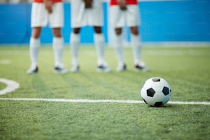 Soccer ball on green football field by dividing line and legs of three players on background