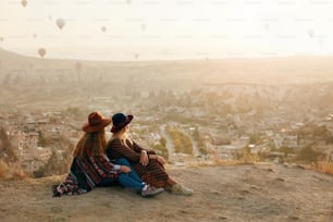 La gente viaja. Mujeres En Sombreros Sentadas En La Colina Disfrutando De Volar Globos Aerostáticos Vista En Capadocia, Turquía. Alta resolución