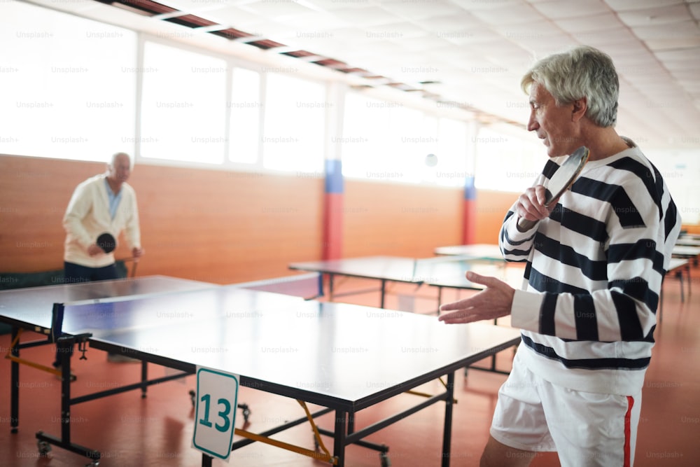 Two active senior men with rackets playing ping pong by large table in the hall