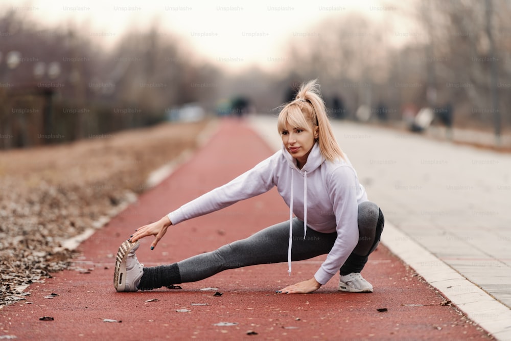 Beautiful blonde woman in sportswear stretching legs before running outdoors.