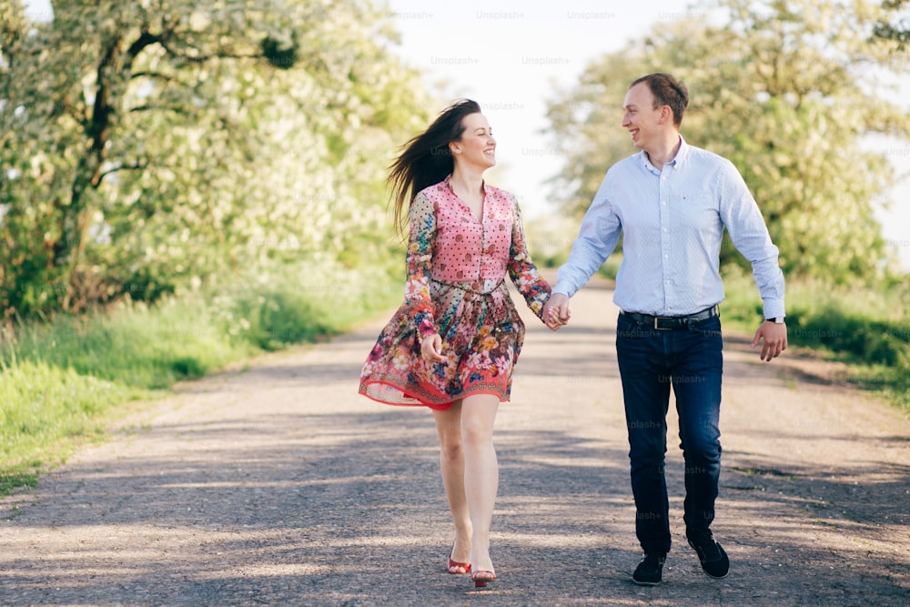 Beautiful young couple holding hands and walking on road in sunshine among spring field and trees. Happy family in love relaxing  in sunlight. Romantic joyful moments. Space for text