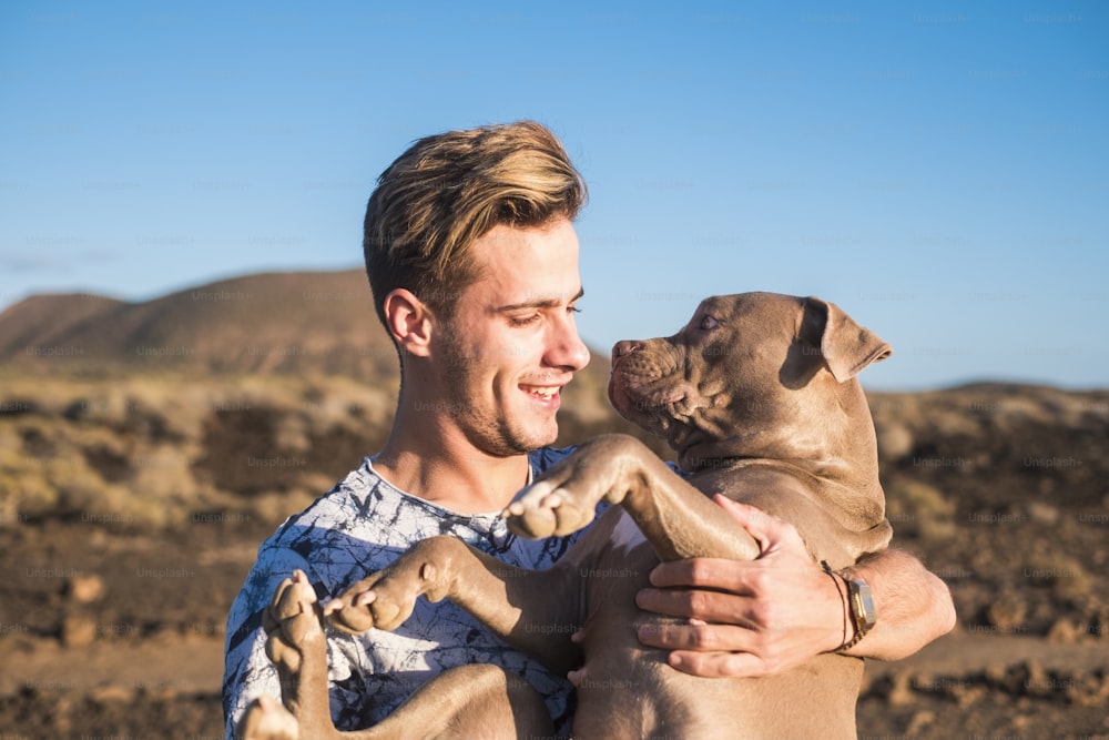 Junger attraktiver blonder Mann Model umarmt mit Liebe und Freundschaft seinen eigenen süßen Welpenhund Amstal in Outdoor-Freizeitaktivitäten und malerischen Wüstenplatz mit Meer im Hintergrund