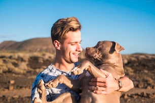 Young attractive blonde man model hug with love and friendship his own cute puppy dog amstal in outdoor leisure activity and scenic desert place with ocean in background