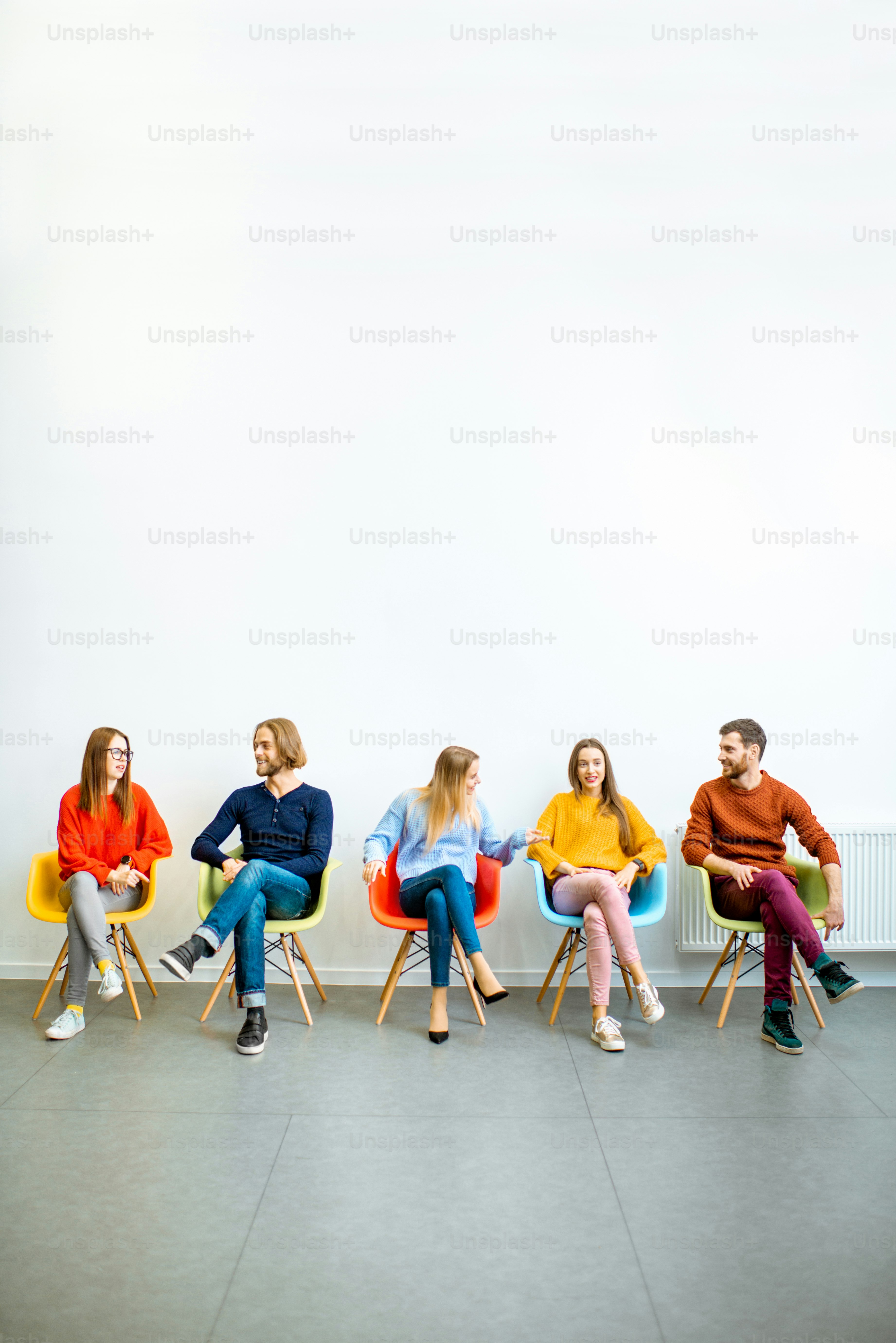 Portrait of a young people dressed casually sitting in a row on the colorful chairs on the white wall background