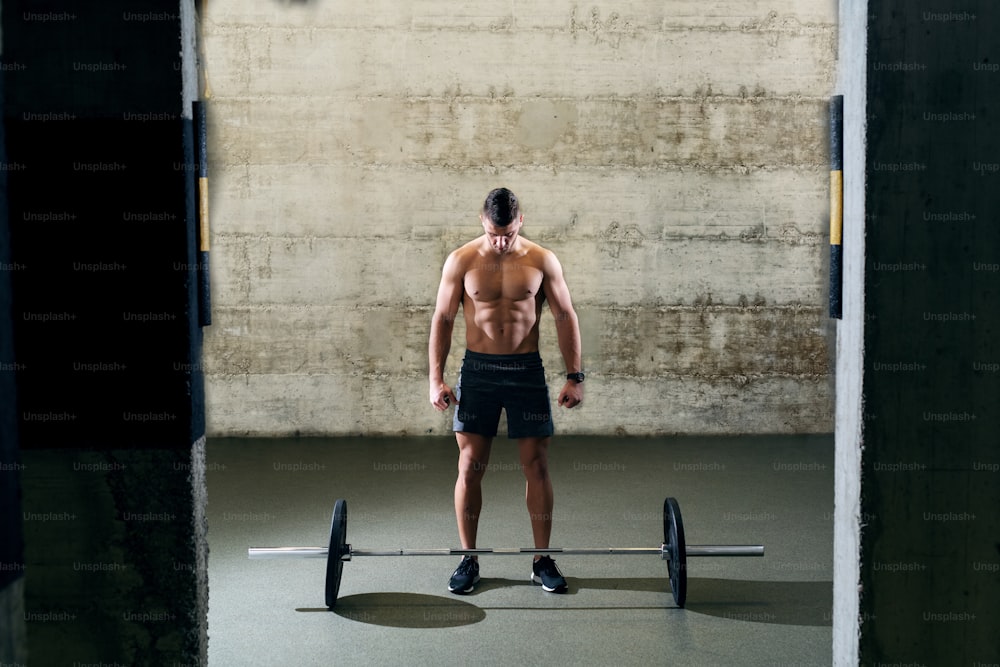 Shirtless Caucasian man in shorts preparing to lift barbell. In background wall.