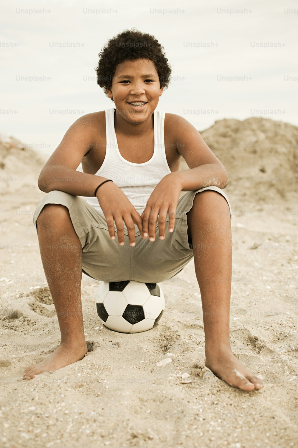 um jovem sentado em cima de uma praia ao lado de uma bola de futebol