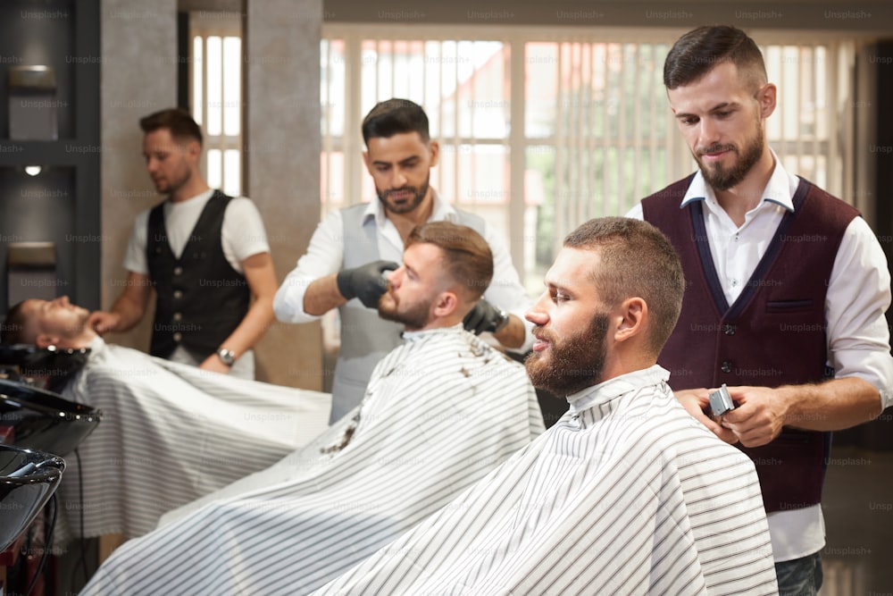 View from side of process of styling hair in vintage barber shop. Brutal handsome barder in uniform keeping trimmer in hands and doing trendy haircut to bearded young man sitting in chair.
