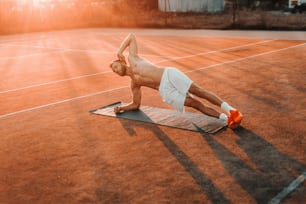 Hombre barbudo musculoso enfocado haciendo planchas laterales en la cancha de tenis por la mañana en verano.