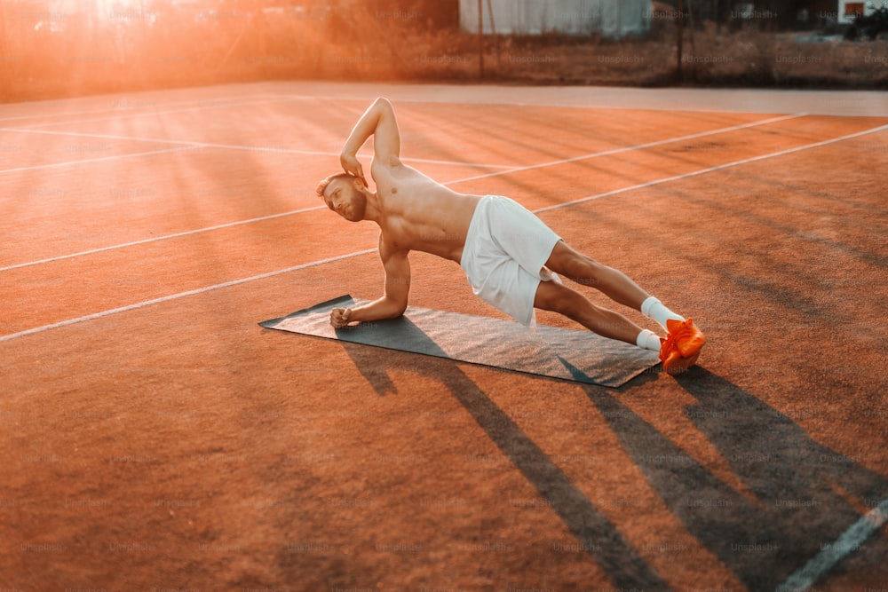 Uomo barbuto muscoloso concentrato che fa plank laterali sul campo da tennis al mattino in estate.