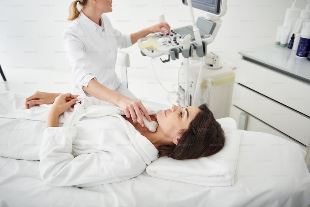 Retrato de vista lateral de una mujer joven atractiva en bata de baño blanca que recibe una exploración ultrasónica de la tiroides