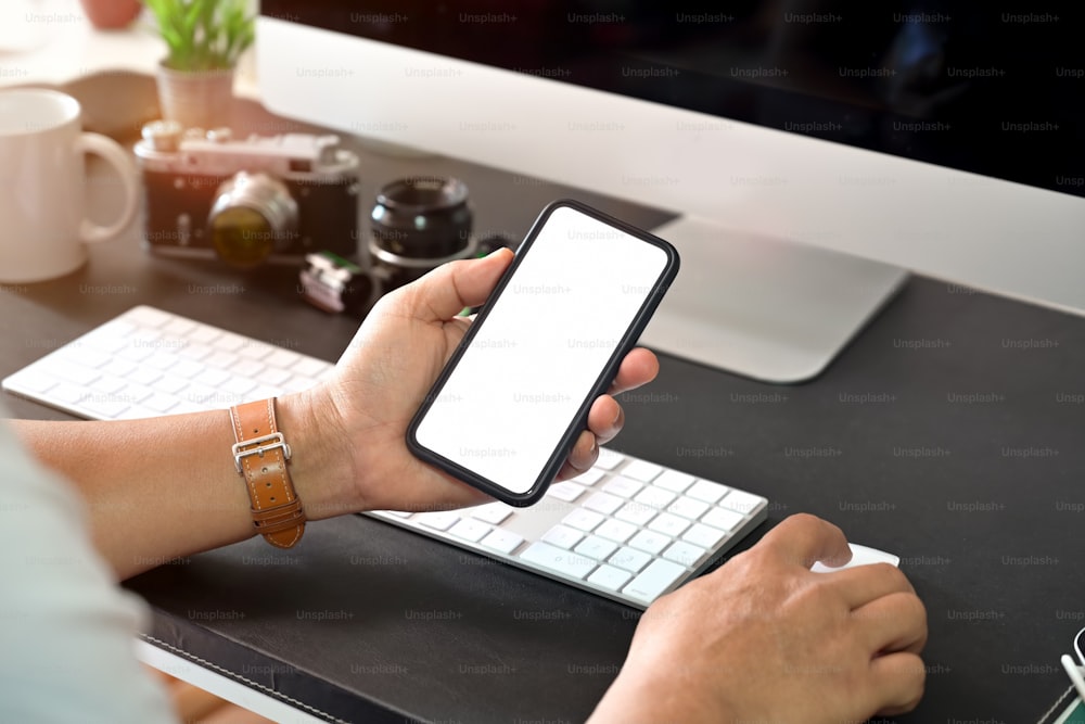 Mão criativa e elegante segurando o telefone móvel de tela em branco enquanto trabalha com o computador de mesa